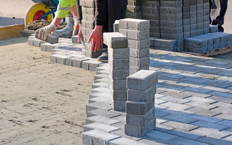 The master in gloves lays paving stones in layers. Garden brick pathway paving by professional paver worker. Laying gray concrete paving slabs on sand foundation base